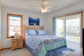 A bedroom with a bed, a nightstand, a lamp, a ceiling fan, a window, and a view of the outside. - Life Is Good: Ocean & Sound Views, Private Pool & Hot Tub, Quick Walk to Beach (Nags Head)