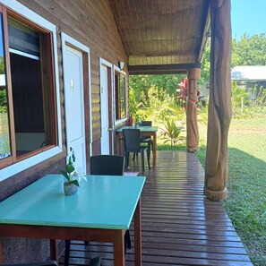 A covered porch with wooden flooring, a table with chairs, and a view of a garden. - Merahi House Cabins I (Isla de Pascua)