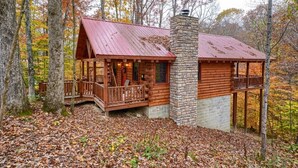 A log cabin with a stone chimney, wooden porch, and a view of the forest.