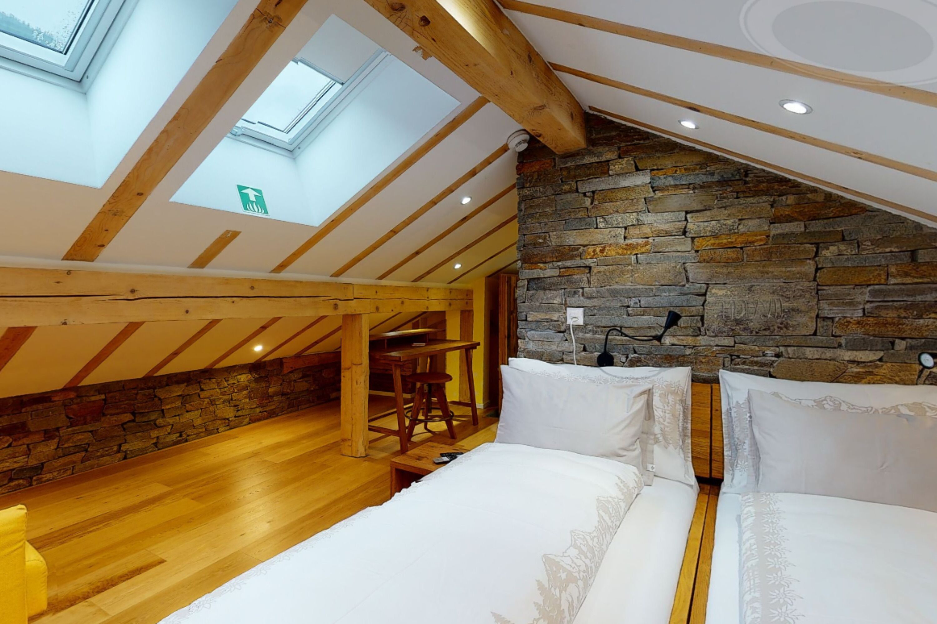 Um quarto com teto inclinado, claraboias, parede de pedra e chão de madeira.