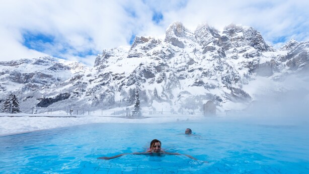 En swimmingpool med kristallklart blått vatten, omgiven av snötäckta berg och en klar himmel.