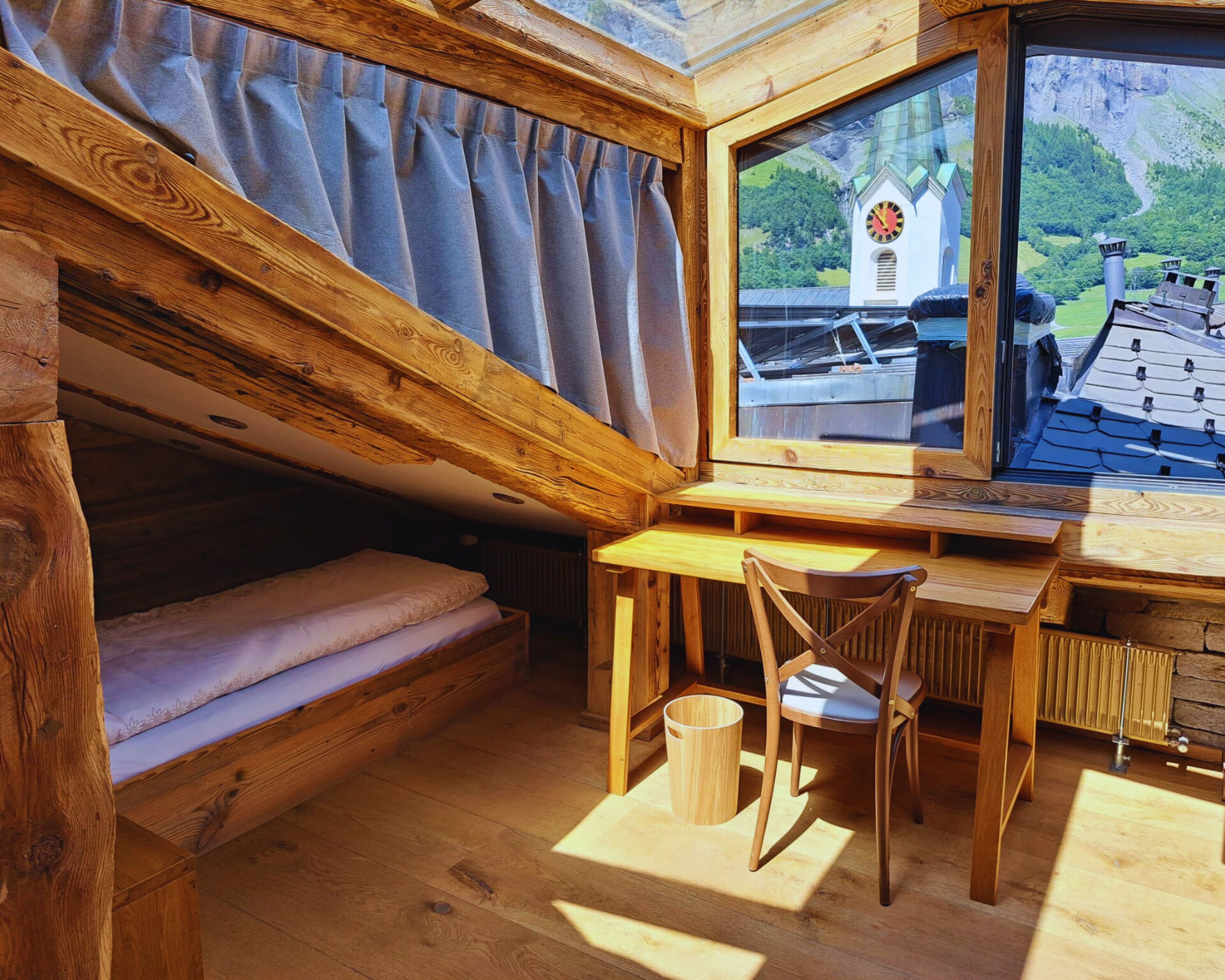 A cozy cabin interior with a wooden bed, a desk with a chair, and a large window overlooking a scenic landscape.