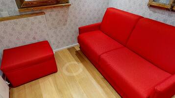 A room with a red sofa and a matching ottoman, wooden flooring, and patterned wallpaper.