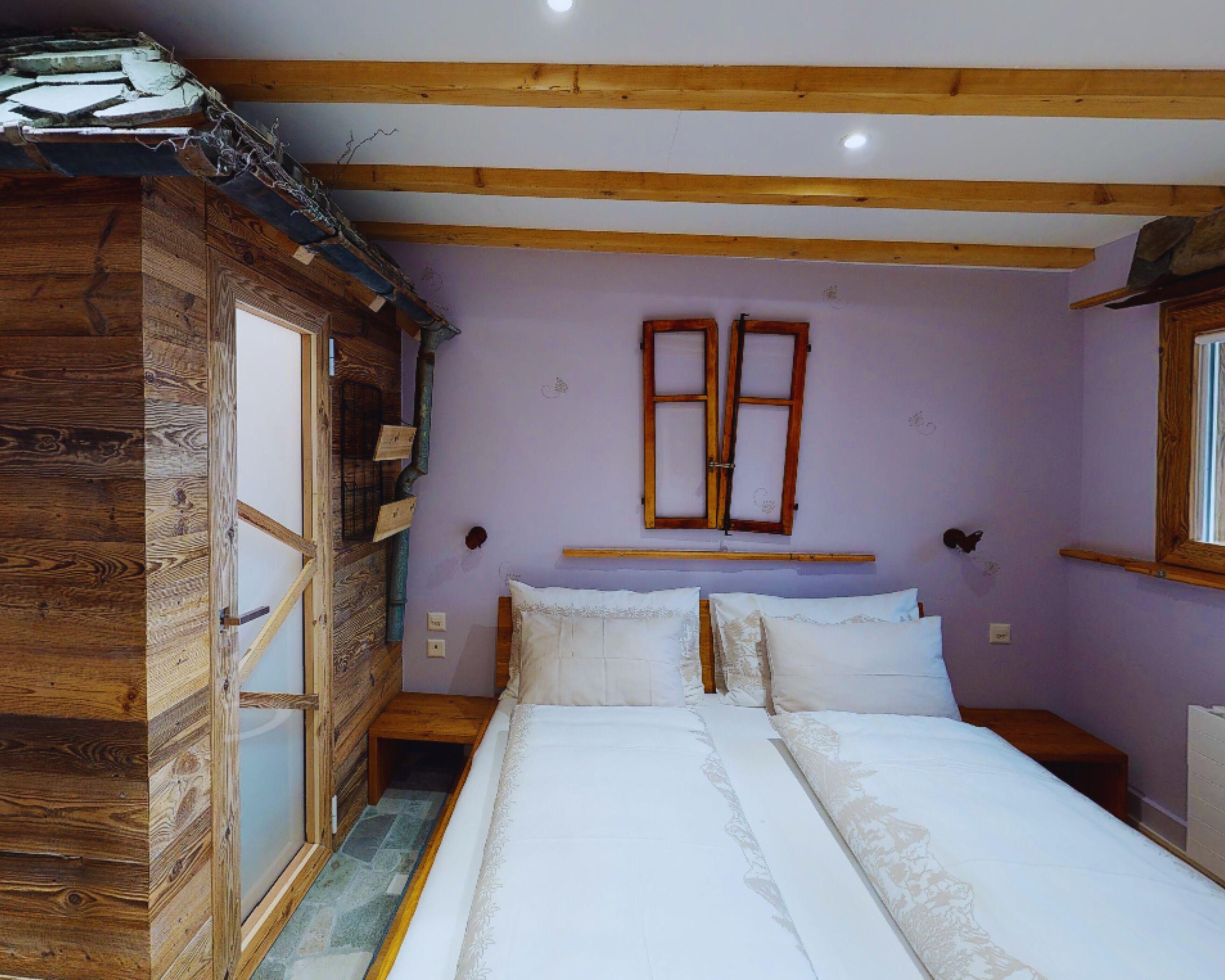A bedroom with a wooden wardrobe, a bed with white linens, a window with wooden shutters, and a small bedside table.
