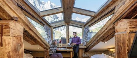 A room with a sloped ceiling and a view of mountains. There is a table with chairs and a person standing.