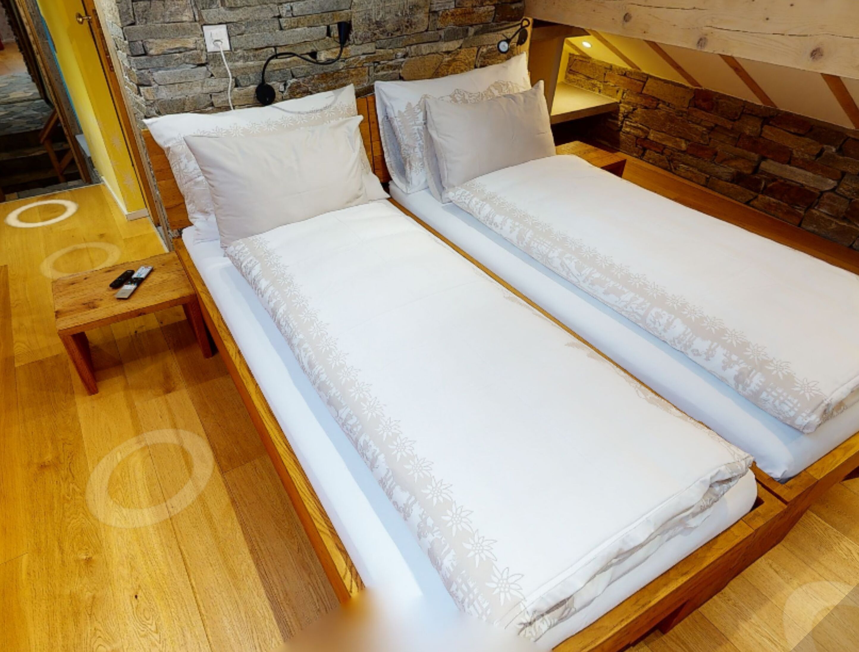 A double bed with white linens in a room with wooden flooring and a stone wall.