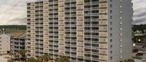 Un hotel de varios pisos con balcones, una playa con sombrillas y un cielo despejado.