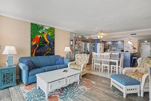 A living room with a blue sofa, white coffee table, and floral armchairs. A colorful parrot painting adorns the wall.