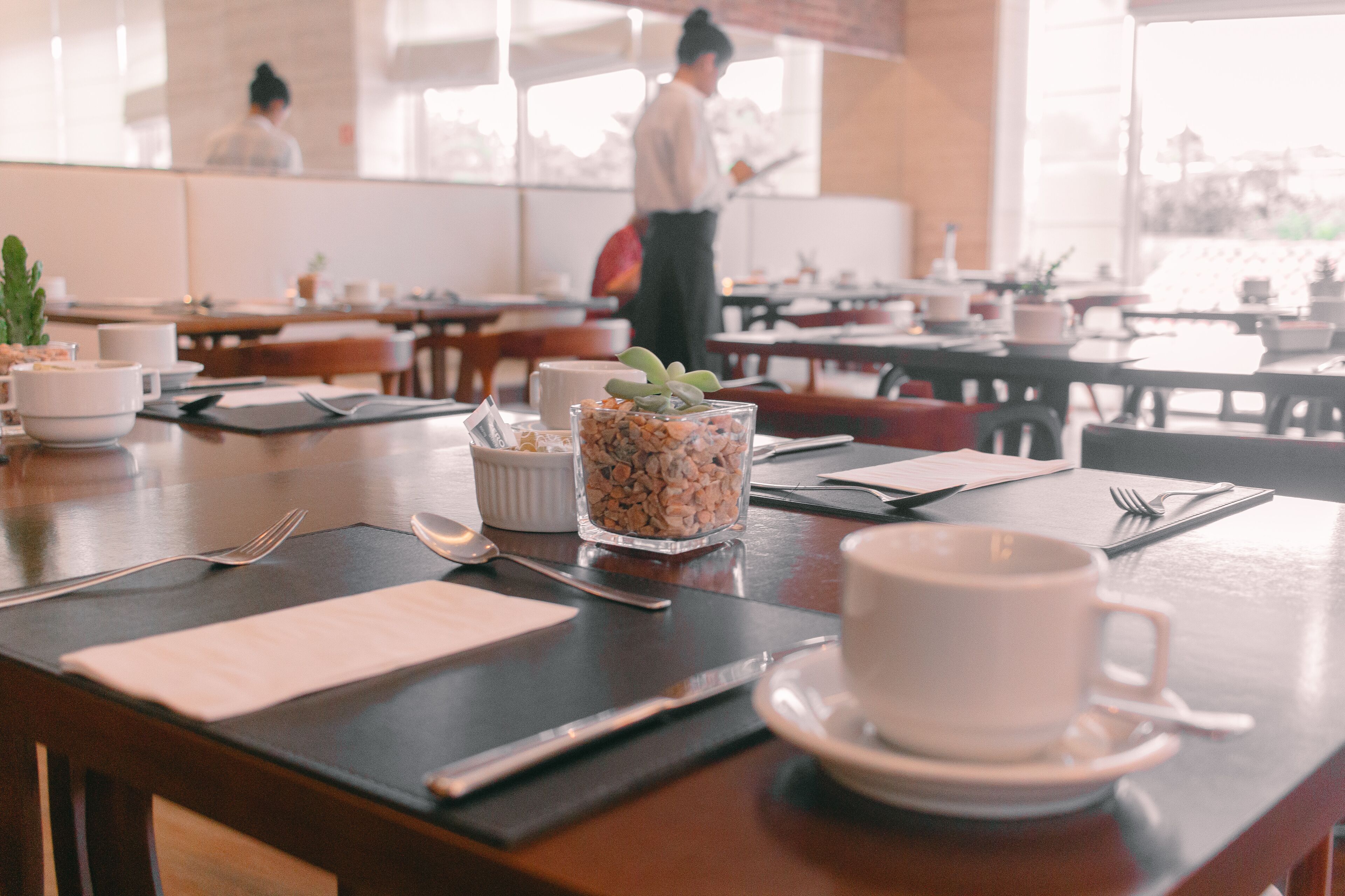 A dining area with tables set for two, featuring white cups, saucers, and cutlery.