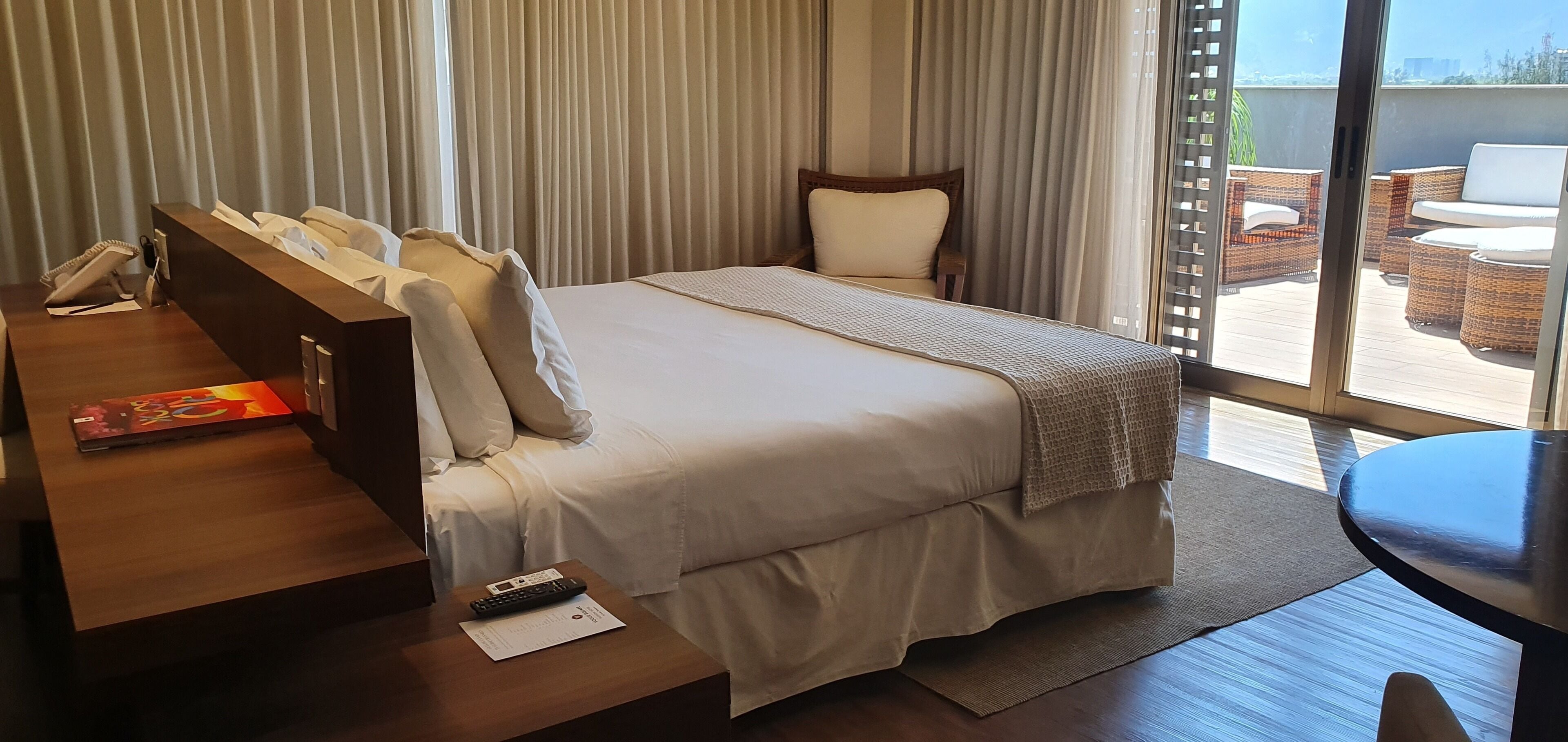 Hotelkamer met een groot bed, een bureau met telefoon, een afstandsbediening en een laptop. Er is een balkon met rieten meubilair.