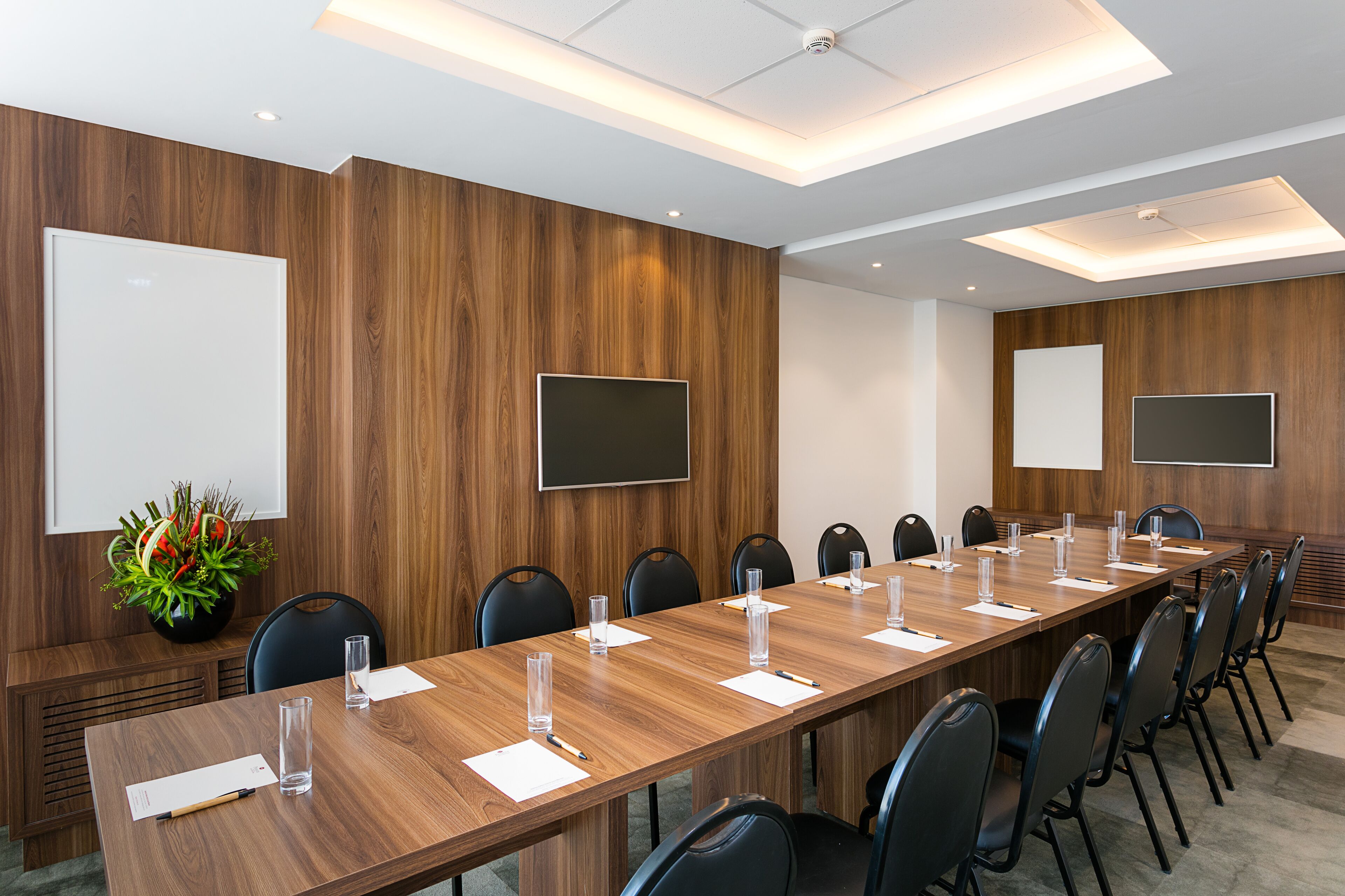 A conference room with a long wooden table, black chairs, white screens, and a potted plant.