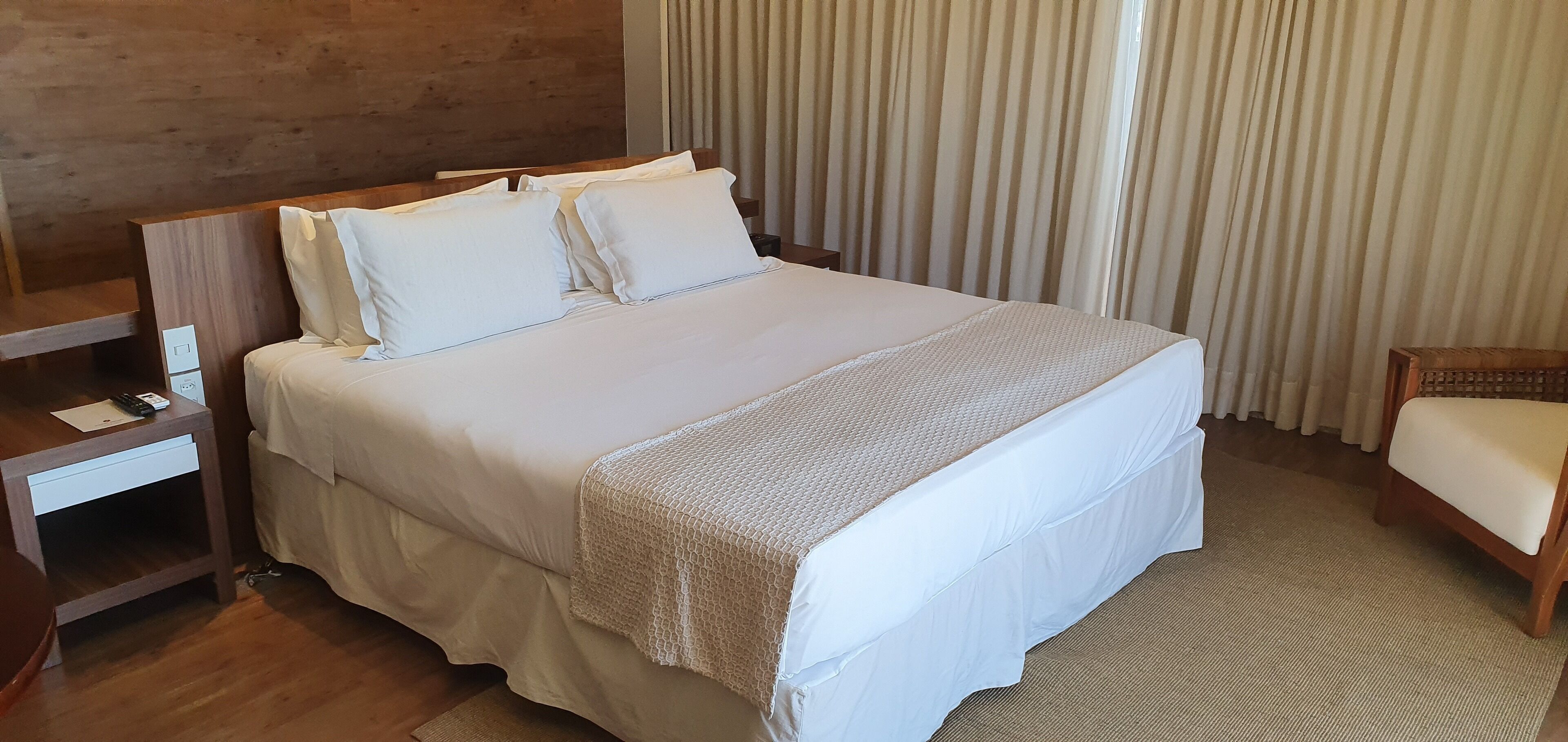 A neatly made bed with white linens and a textured blanket, a wooden nightstand with a remote control, a wooden chair with a cushion, and a window with vertical blinds.