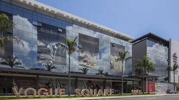 Um edifício moderno com grandes janelas de vidro e faixas publicitárias com os dizeres 'Academia', 'Gourmet' e 'Hotel'.