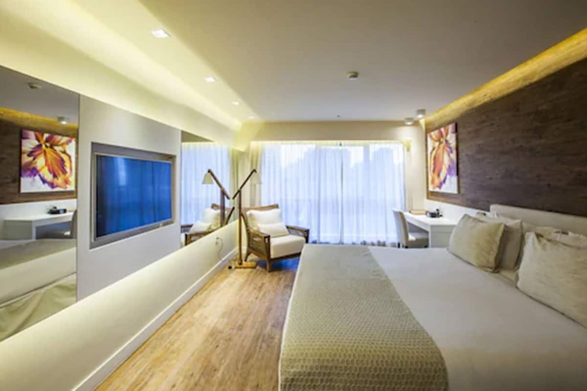 Ett modernt hotellrum med en stor säng, ett skrivbord, en stol, en TV och ett stort fönster med gardiner.