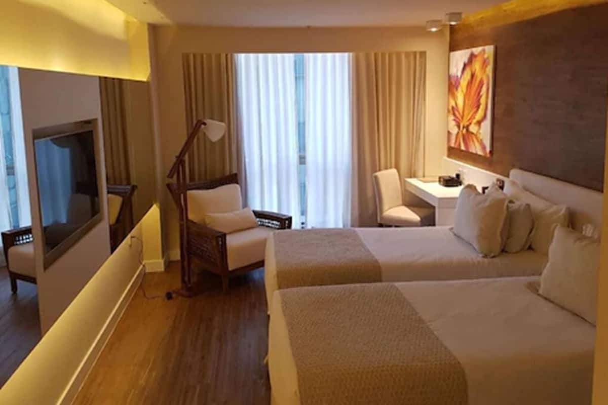 Ett hotellrum med två sängar, ett skrivbord, en stol, en TV och ett stort fönster med gardiner.