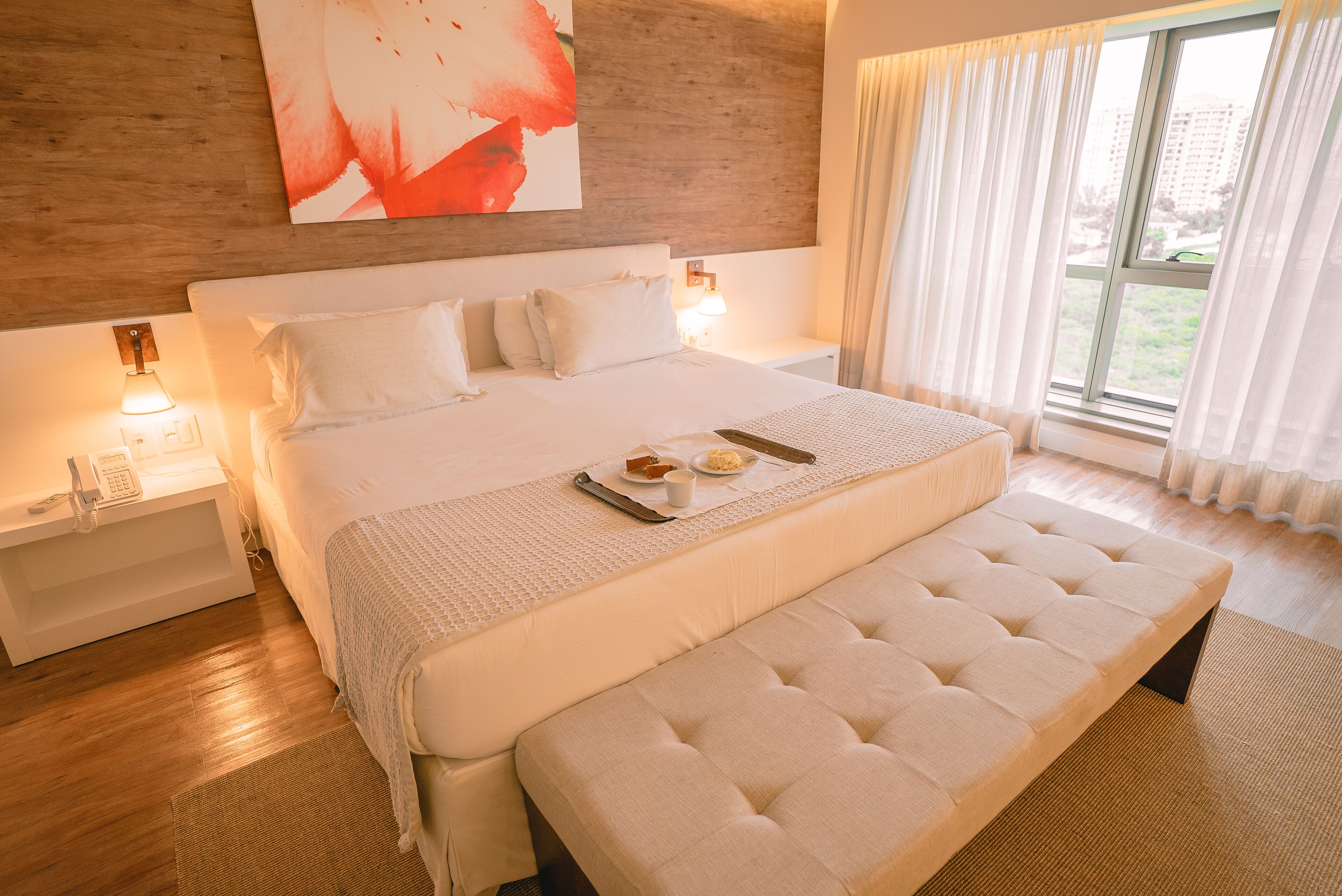 Un dormitorio moderno con una cama grande, mesitas de noche, un banco y vistas al exterior a través de una ventana.