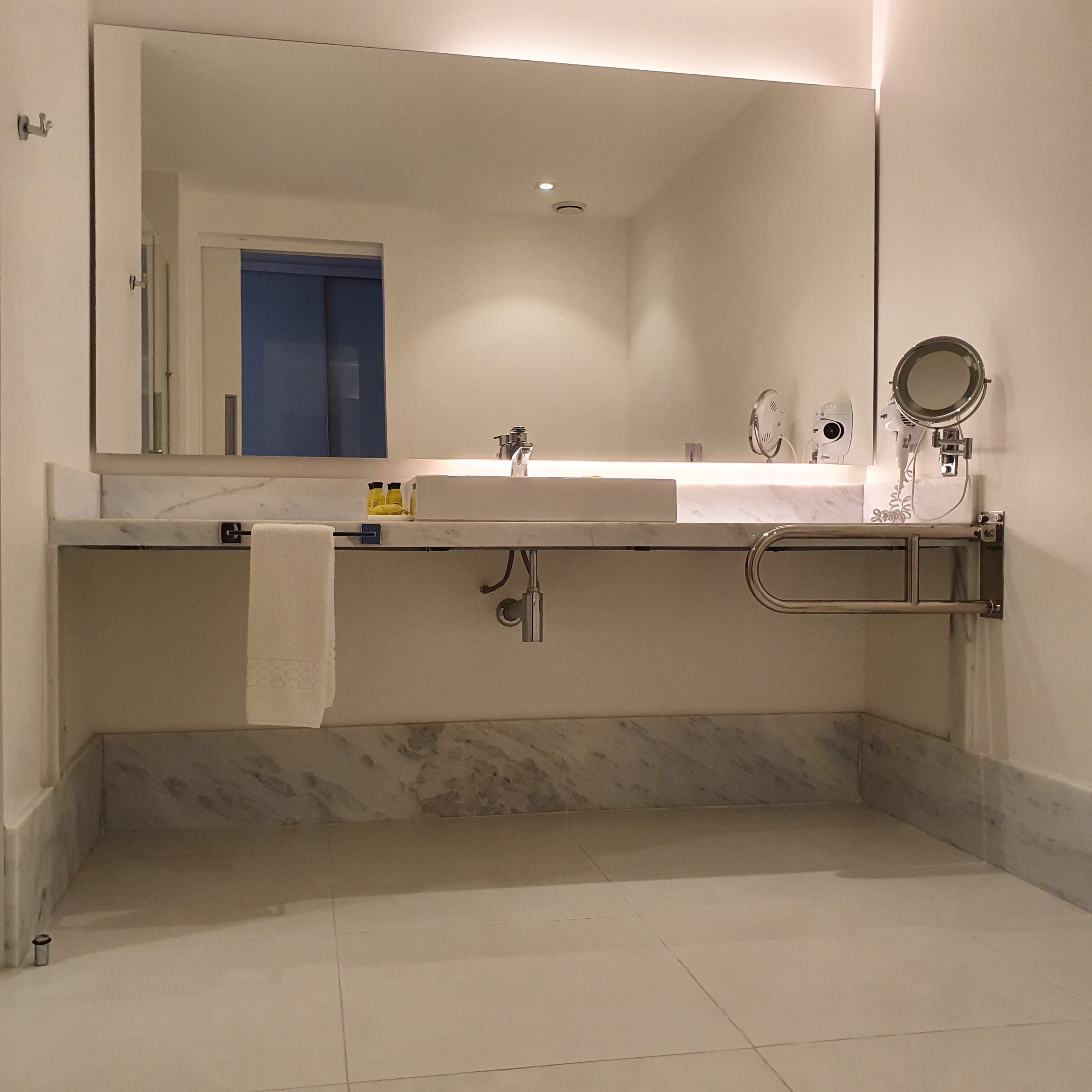 A modern bathroom with a large mirror, two sinks, a towel rack, and a handheld mirror.