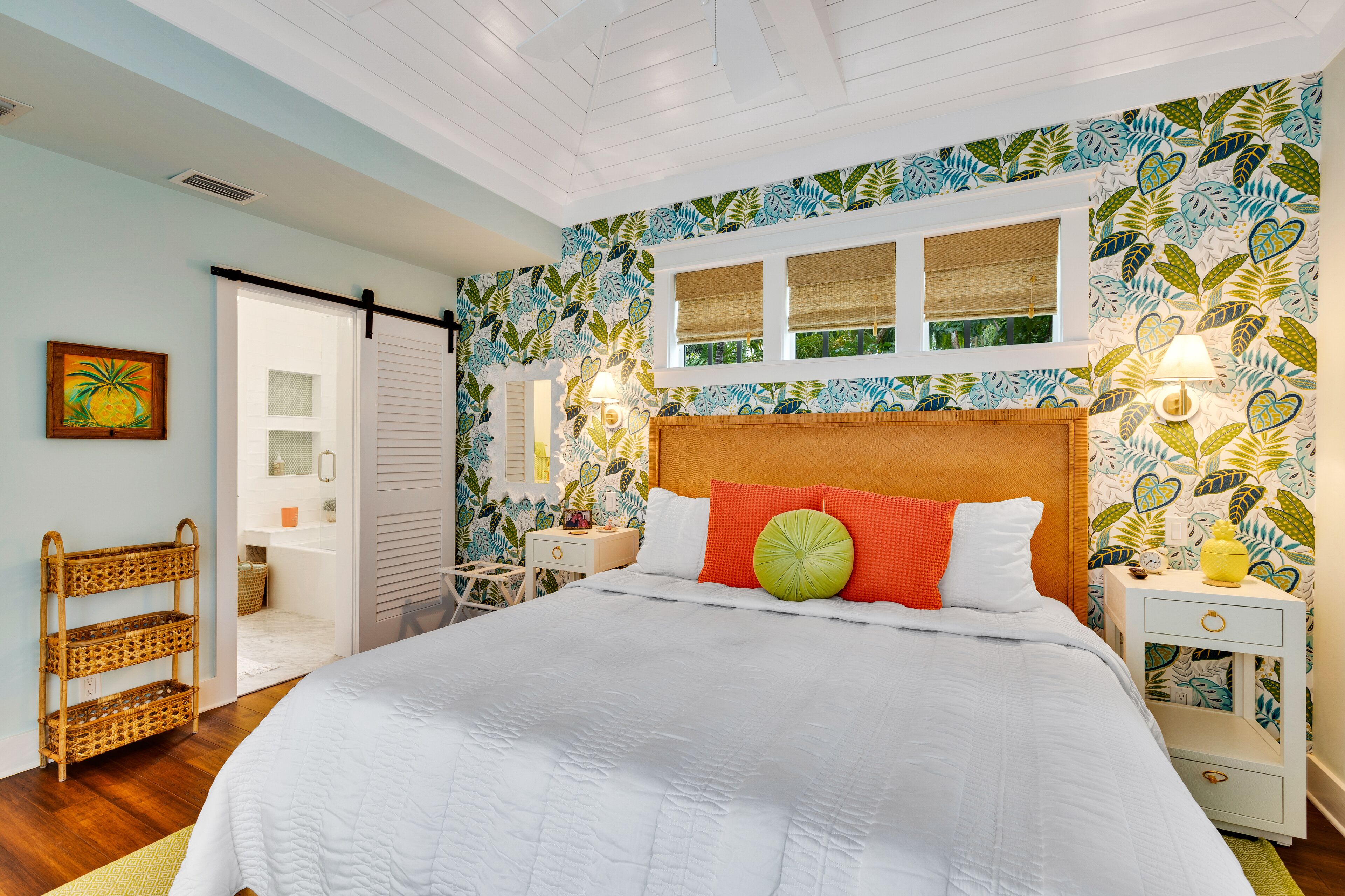 A bedroom with a large bed, a nightstand, a wicker chair, a window, and a door leading to a bathroom.