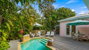 Eine Holzterrasse mit Swimmingpool, umgeben von üppiger Vegetation und einem pinkfarbenen Gebäude.