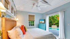 Ein Schlafzimmer mit Bett, Nachttisch, Kommode, Deckenventilator, Fenster mit Blick auf einen Pool und einer tropisch gestalteten Wand.