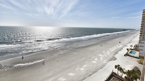 A beach with a long pier extending into the ocean, a sandy beach, and a building with a pool on the right.