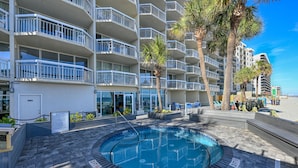 A hotel with a circular pool surrounded by a brick patio, palm trees, and a beach view.