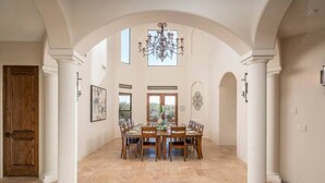 Une salle à manger spacieuse avec un plafond haut, un lustre et une vue sur l’extérieur par les fenêtres.