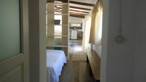 Passadís estret amb una banca de fusta, un llit amb roba de llit blanca i una zona de cuina amb nevera i armaris.