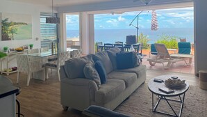 Een moderne woonkamer met een bank, eettafel en stoelen. Een groot raam biedt uitzicht op de oceaan.