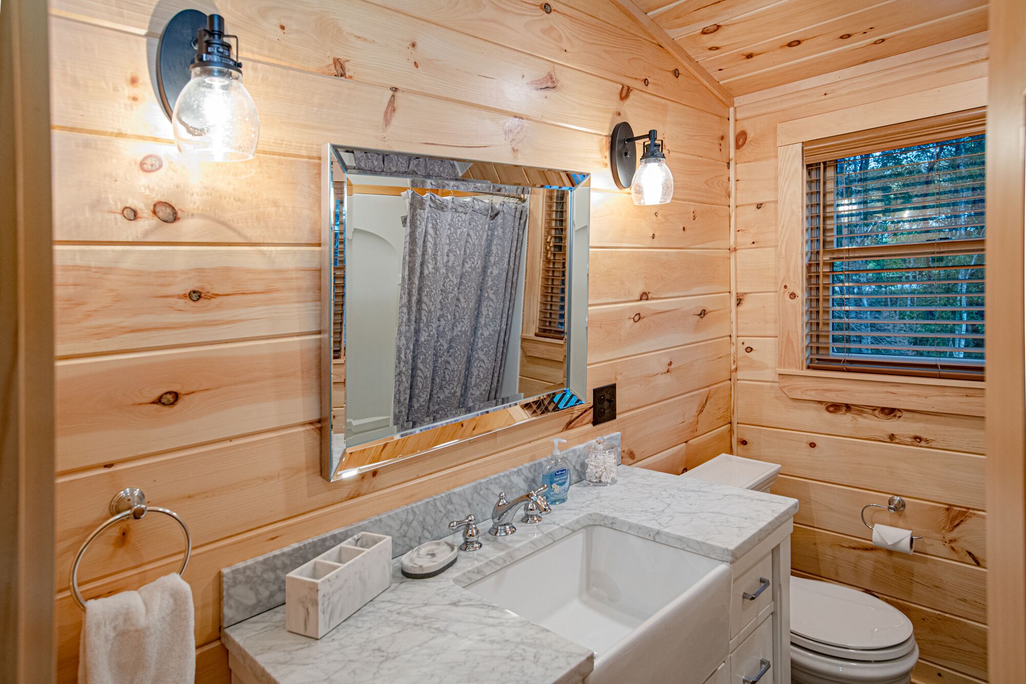 Bagno di una cabina in legno con uno specchio grande, un lavabo con piano in marmo, un water e una finestra con persiane.