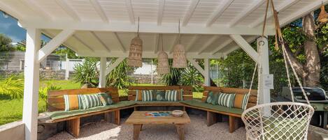 A covered outdoor seating area with a wooden bench and a hanging chair, surrounded by greenery.