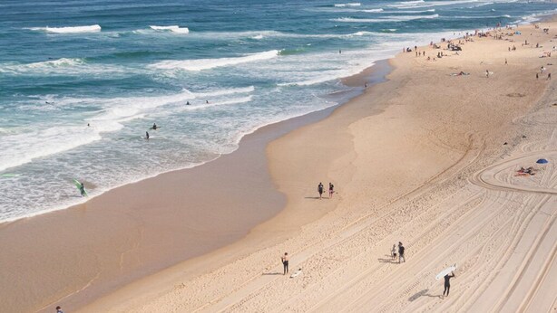 Una amplia playa de arena con olas rompiendo en la orilla, gente caminando y algunos surfistas en el agua.