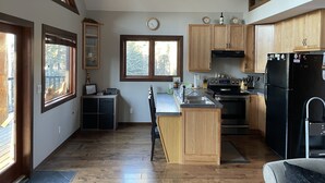 A kitchen with wooden cabinets, a black refrigerator, and a window overlooking a deck. - Rocky Mountain Cottage (Nordegg)