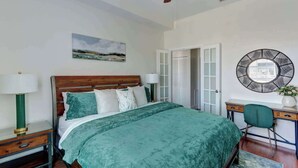 Un dormitorio con cama, escritorio, silla, ventilador de techo, puerta y espejo montado en la pared.