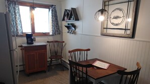 A compact kitchen with a refrigerator, a coffee maker, and a dining table with chairs. A window with curtains is visible, and a sign on the wall reads "Fresh Brewed Coffee Served Daily."
