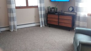 Una sala de estar con una televisión mostrando "Catch me if you can", un aparador de madera, un reloj y una ventana con cortinas.