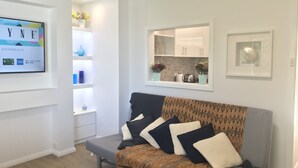 A modern living room with a grey sofa, patterned throw, and decorative pillows. A white wall-mounted TV displays a logo and text. A kitchen area is visible through an open doorway, featuring cabinets and a countertop.