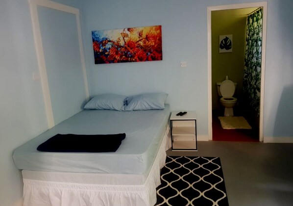 A bedroom with a bed, a nightstand, a painting, and a bathroom visible through an open door.