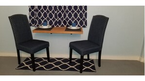 Two dark blue upholstered chairs with metal studs, a wooden shelf with two white plates and blue cups, and a black and white patterned rug on a light blue wall with a geometric patterned curtain.