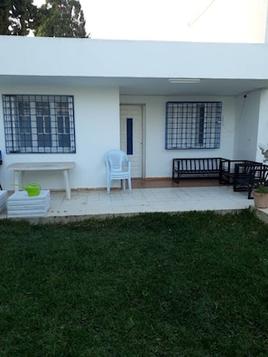 A white building with a covered patio area. There are two windows with blue grills, a white door, and a small white chair. - Villa LARIBI seaside in Nabeul. (nabeul)