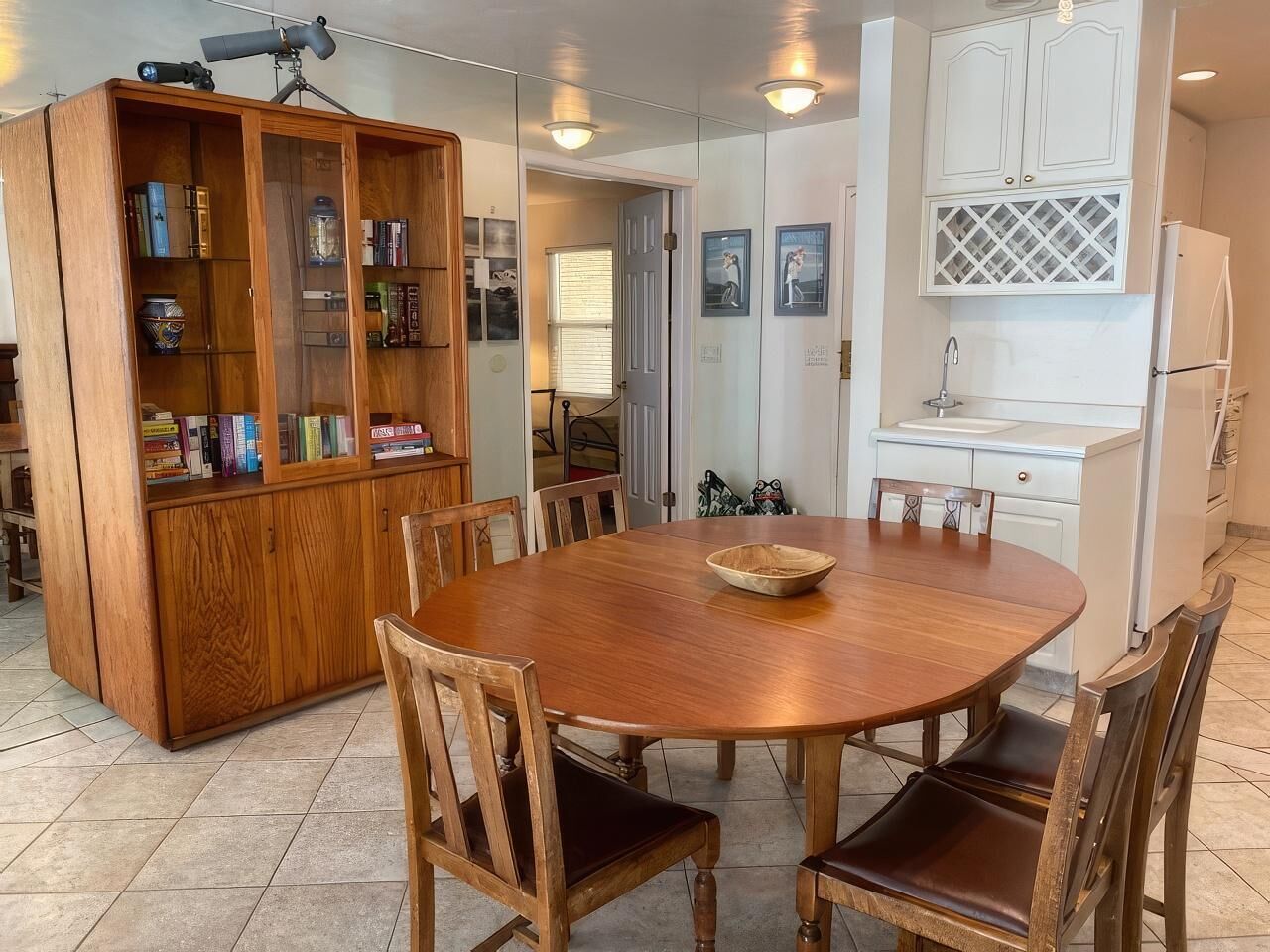 A dining area with a wooden table and chairs, a wooden cabinet with books, and a kitchen area with white cabinets and a sink.