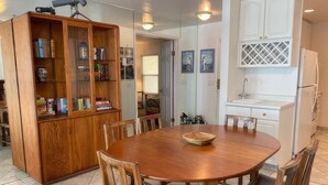 A dining area with a wooden table and chairs, a wooden cabinet with books, and a kitchen area with white cabinets and a sink. - See The Sea 2-Ocean Front, Dining on Patio, 2 bd! (San Diego)