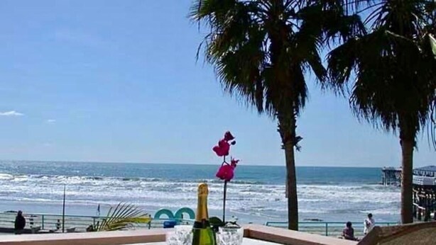 A table with a bottle and glasses, overlooking the ocean with palm trees and a pier in the background. - See The Sea 2-Ocean Front, Dining on Patio, 2 bd! (San Diego)