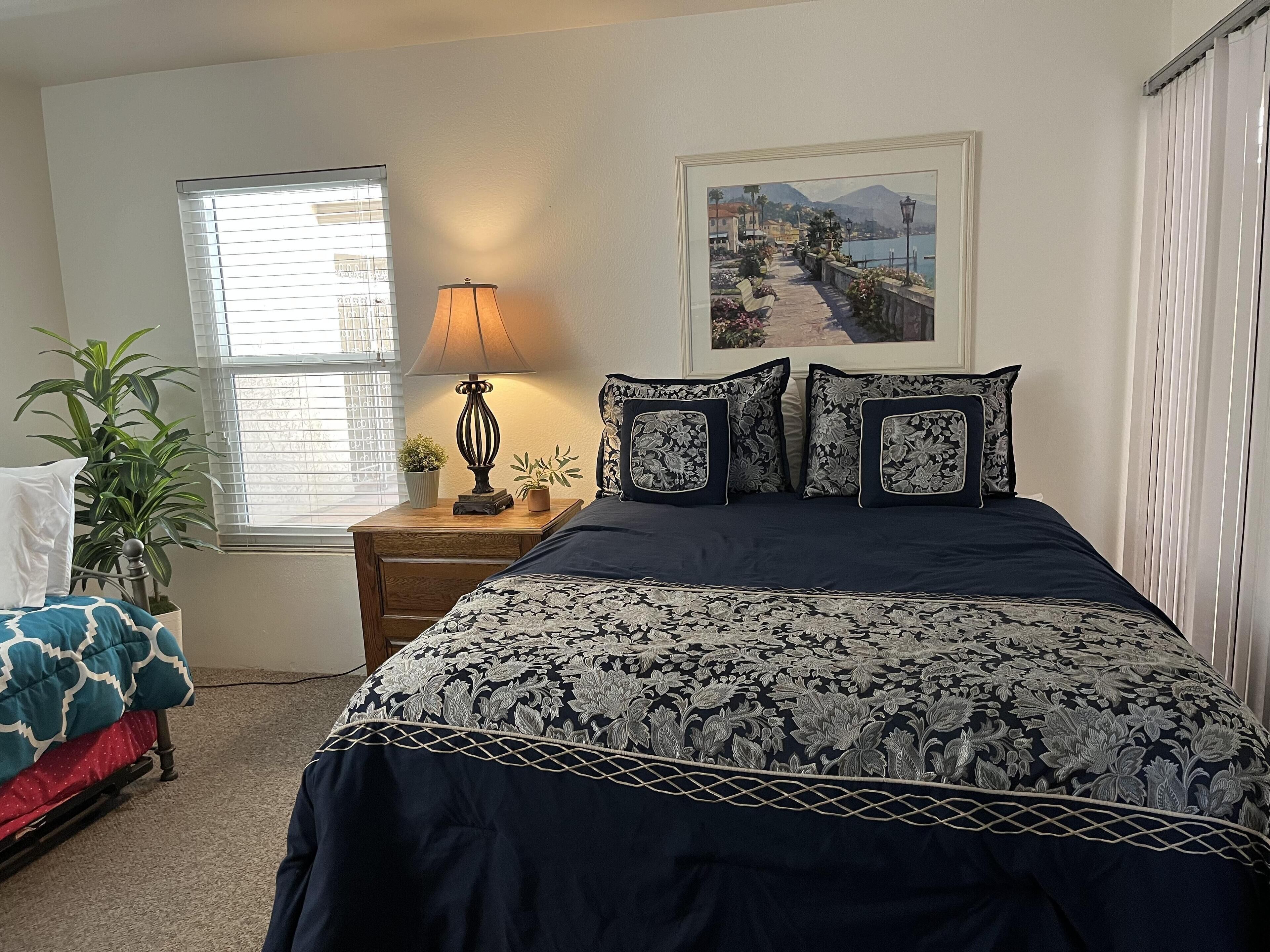 A bedroom with a bed, a nightstand with a lamp, a potted plant, and a window with blinds.