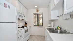 A compact kitchen with white cabinets, a stainless steel sink, a window with blinds, and a clock on the wall. - See The Sea 2-Ocean Front, Dining on Patio, 2 bd! (San Diego)