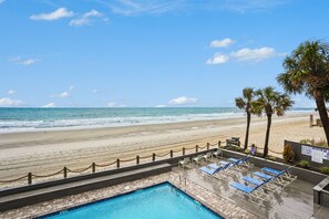 A pool area with a view of the beach and ocean, lounge chairs, and a wooden deck.