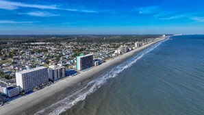 An aerial view of a coastal city with a long beachfront, high-rise hotels, and a clear blue sky.
