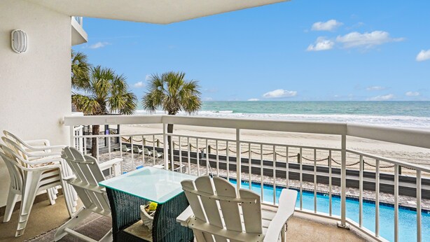 A balcony with a glass table and white chairs, overlooking a beach and a pool.