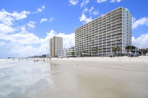 A beachfront hotel with multiple floors, a sandy beach, and palm trees.