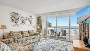 A living room with a sofa, a coffee table, a lamp, and a large window with a view of the beach.